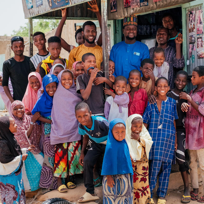 Nigerian refugees gathered in the shadow of a small house and smiling.