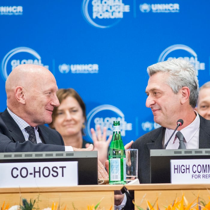 Two men shake hands on a podium behind the signs 'Co-host' and 'High Commissioner for refugees'.