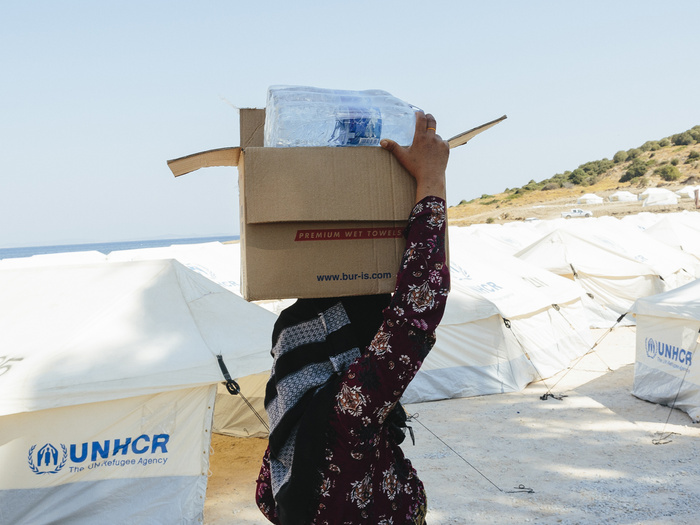 A woman carrying a cardboard box on her head in front of white tents with the UNHCR logo.