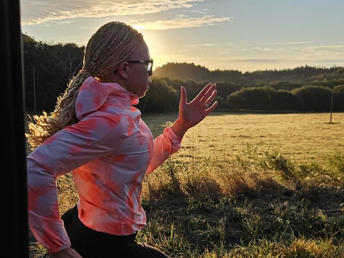 A woman with long, tied back braids runs through the countryside, the sun behind her