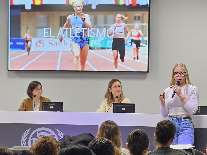 A woman addresses a seated crowd from the front of the room, with two seated people with computer screens and microphones behind her. A large screen on the wall shows her running on an athletics track, with the words El Atletismo overlayed.