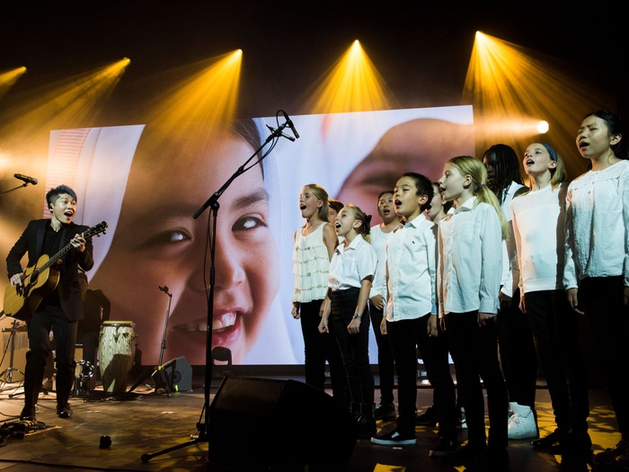 MIYAVI on stage performing with his guitar, a choir of children in white shirts and black pants to his right.