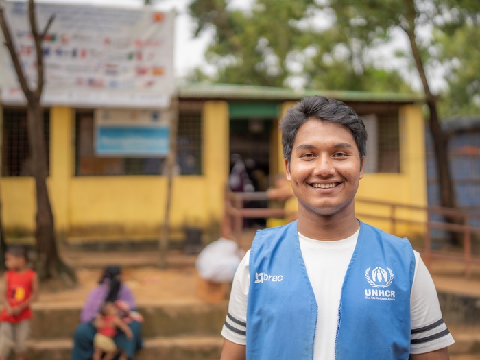 A man in a UNHCR vest smiles proudly in front of a building