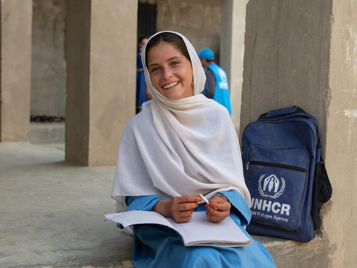 A primary-aged Afghan girl smiles at the camera with a book on her lap and a UNHCR backpack beside her.