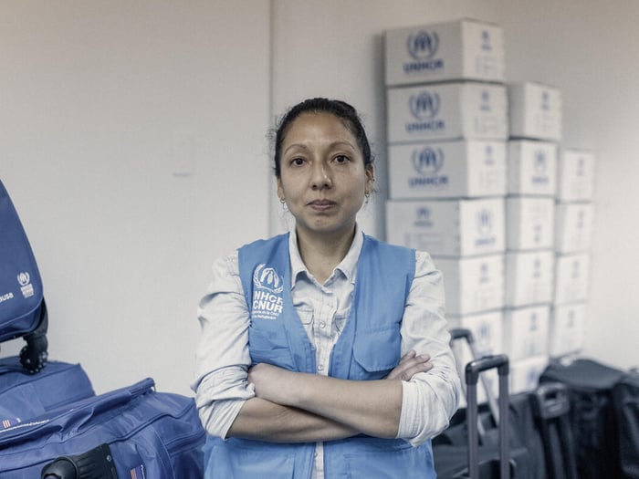 A UNHCR protection officer poses for a photo in a supply closet containing boxes, suitcases and bags of supplies.