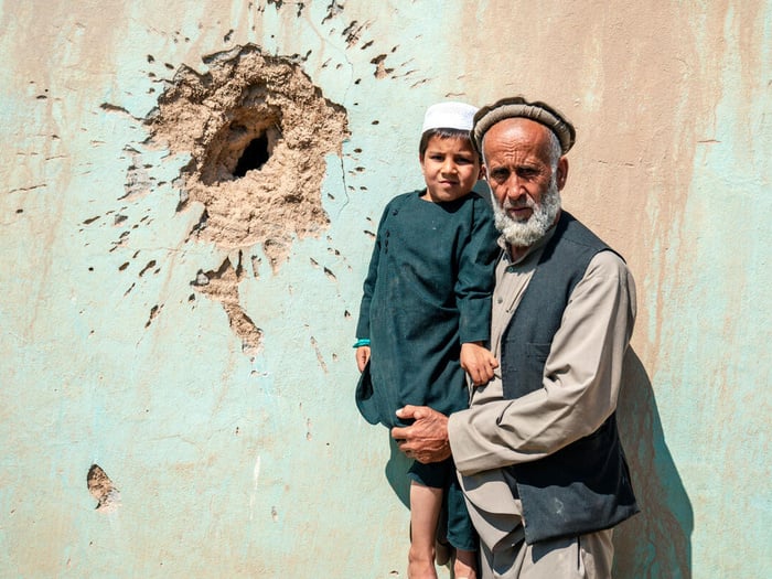 A man holds a young boy in his arms, standing in front of a wall damaged by conflict.