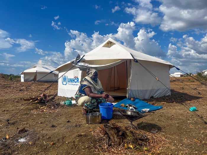 A smiling woman squats in front of a UNHCR tent pitched on dirt, with blue sky and white clouds behind.