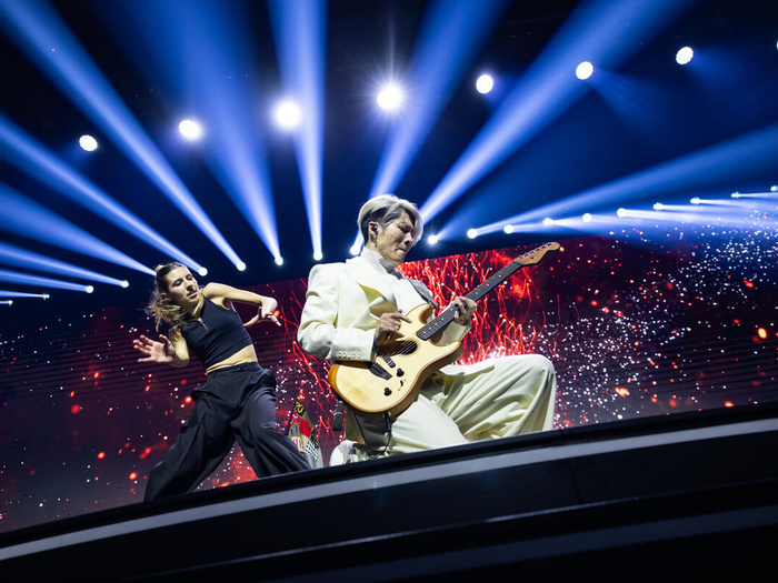 MIYAVI on stage on his knees playing a guitar, a dancer and bright lights behind him