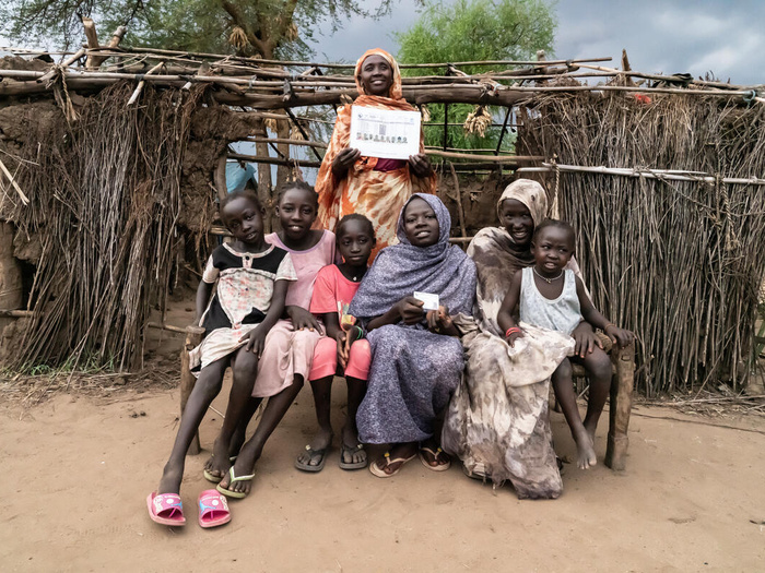 A family is pictured, smiling, in front of a shelter, with two members displaying their IDs.