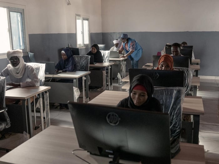 Students sit in rows behind desks with computer screens, while a UNHCR staff member stands leaning over one student's desk.