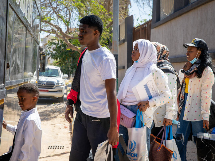 An 18-year-old man from Sudan boards a bus with a young child in front of him and other adults behind him. Several of them are carrying UNHCR-branded bags.