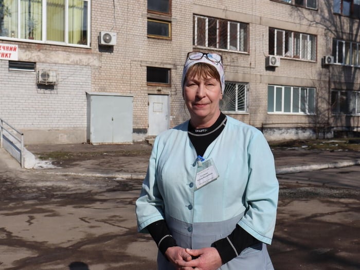 A woman stands outside a building, wearing a nurse's uniform
