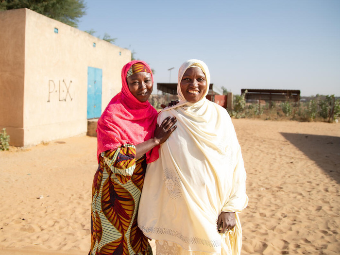 Two friends, A Nigerian refugee woman and an internally displaced woman from Niger, stand together smiling, their arms around each other, in their settlement.