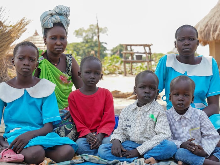 A family of 6 siblings sit together on a mat on the ground outside, some shelters with straw rooves behind them.