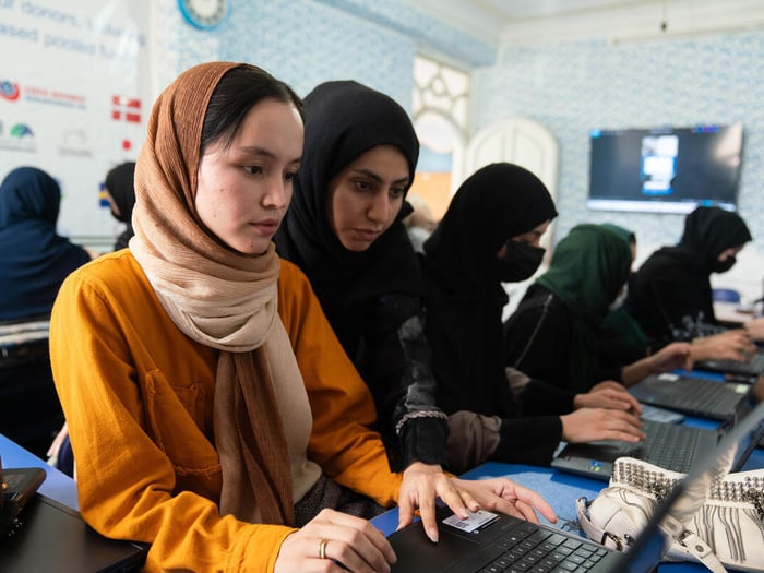Two girls stare intently at a laptop, with other students beside and behind them also working on laptops