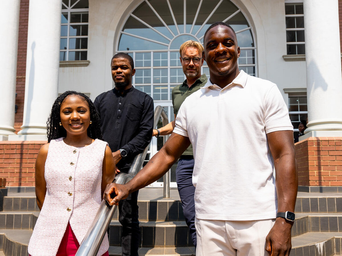 Emmanuel Tshituka, a 25-year-old man, is pictured smiling on a set of stairs, a woman beside him and two men behind them.