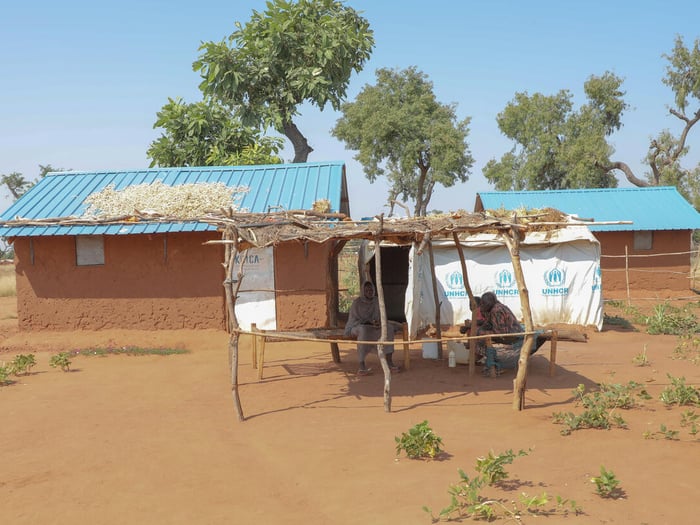 A Sudanese family sit together in the shade outside their shelter, which shows branding from UNHCR and KOICA.