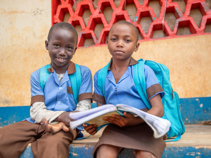 Two Central African refugee children sit outside in school uniform, holding open a book together.