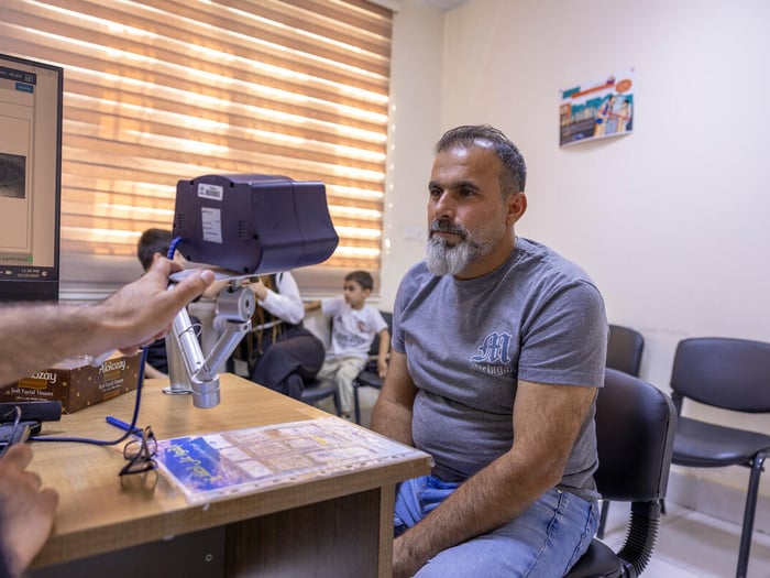 A male Syrian refugee has his eyes scanned as he sits in a room with the curtain down, while two children and another adult sit behind him.