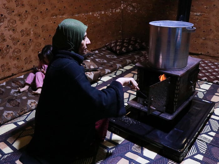 A Syrian woman crouches in front of an indoor wood stove, with a small child holding a mobile phone behind her.