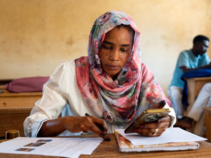 A Sudanese woman sits at a desk, concentrating on a phone, book and papers in front of her.