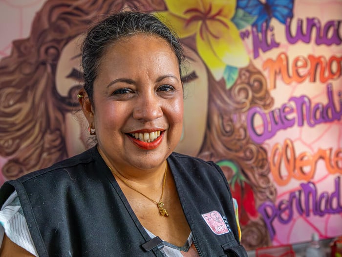 A woman pictured smiling in front of a colourful mural. It reads It reads: “Ni una menos. Que nadie llore por nadie” (“Not one less. Let no one mourn another”)