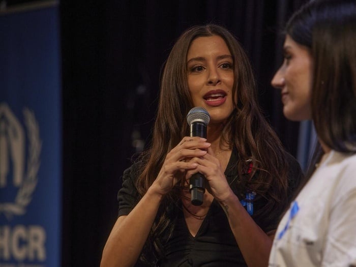 Marina Satti speaks into a microphone while looking at someone beside her, a UNHCR banner in the background
