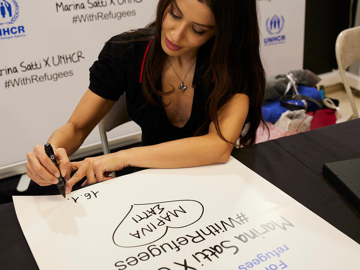 Marina signs a large poster with the UNHCR logo which reads "For a world where refugees are welcomed. Marina Satti X UNHCR. #WithRefugees"