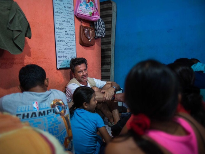 Osvaldo Laport sits on the floor with refugees and asylum-seekers in a room with coloured walls, bags and a framed picture handing behind him.