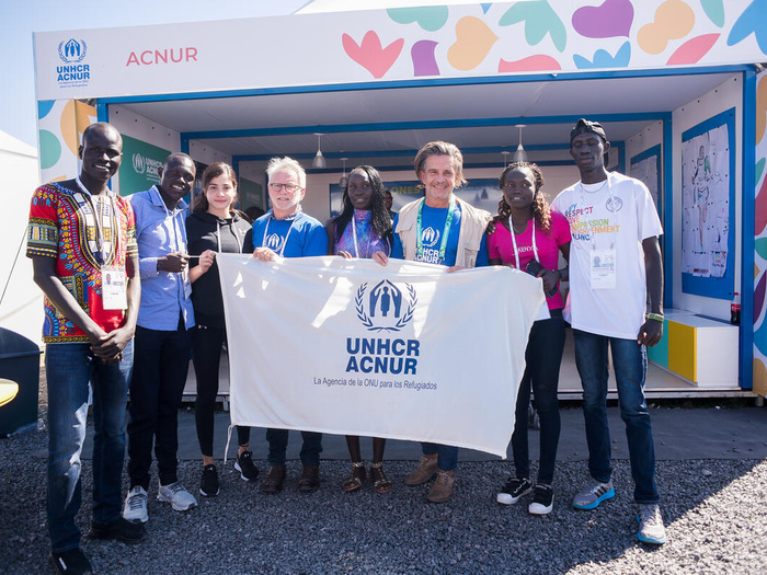 Two UNHCR representatives and members of the Refugee Olympic Team hold up a UNHCR/ACNUR banner