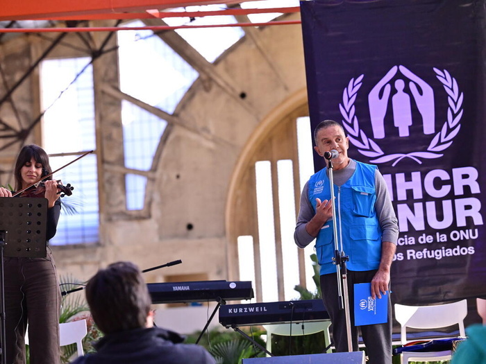 Osvaldo Laport speaks on a stage in a UNHCR vest, a UNHCR banner behind him, and a violinist playing beside him, some keyboards between them