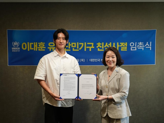 Daehoon Lee and Sae-Ryo Kim pose side by side, holding two signed papers in a display folder. A banner in Korean is behind them.