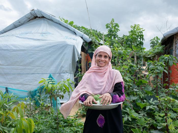 A woman holding a plate of fresh vegetables while standing in front of a garden in a refugee camp. 