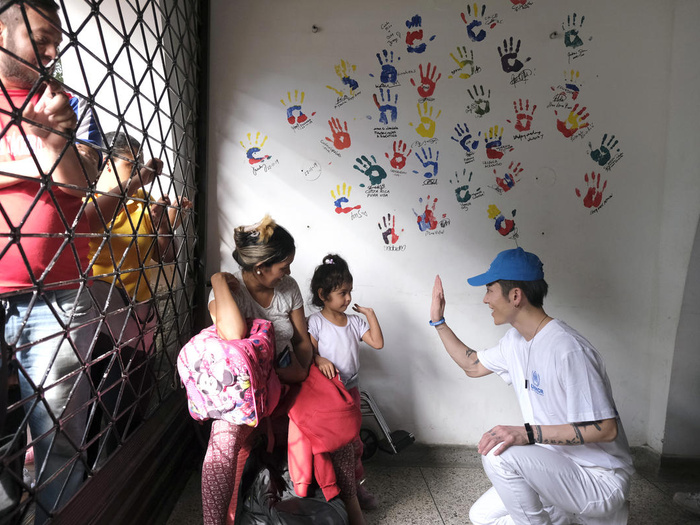 MIYAVI high-fives a young child with a wall of painted hand prints behind them. Others stand nearby behind a fence.