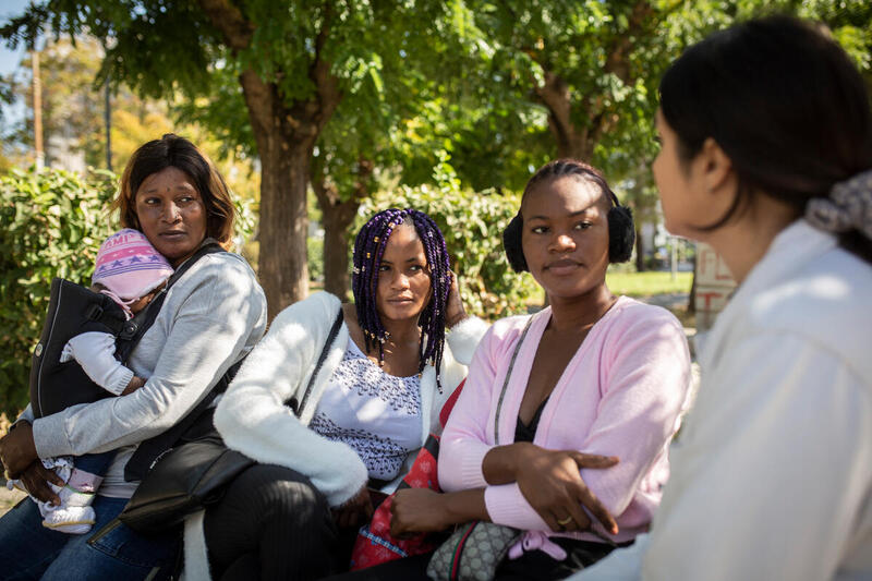 Three refugee women, including one holding an infant, sit on a bench and are listening to a woman working for UNHCR.