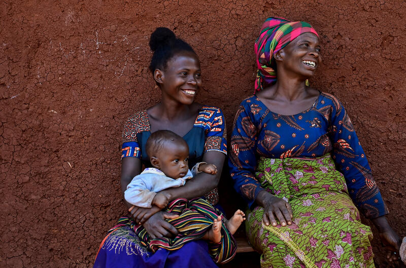 Two smiling women sit on a bench, one holding a baby.