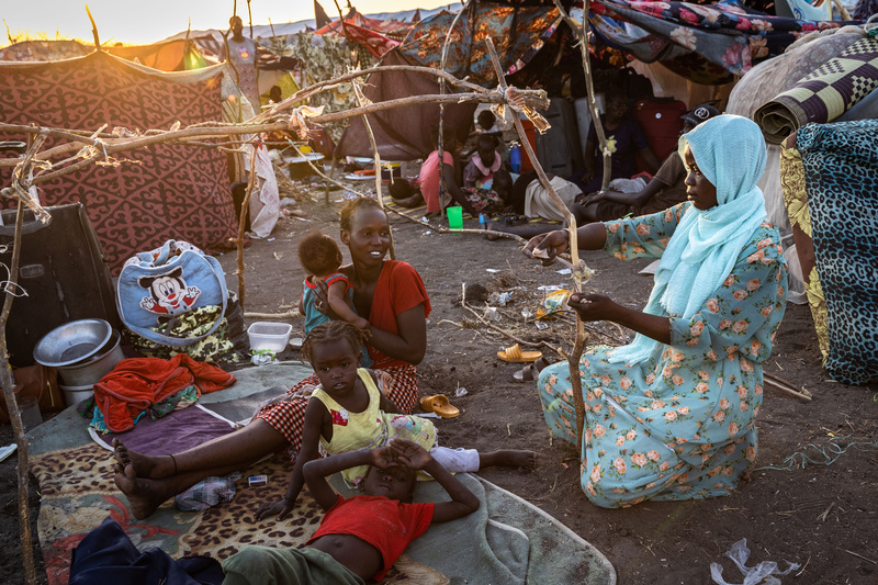 Une femme assise par terre avec ses trois enfants regarde une autre femme attacher des bâtons pour un abri.
