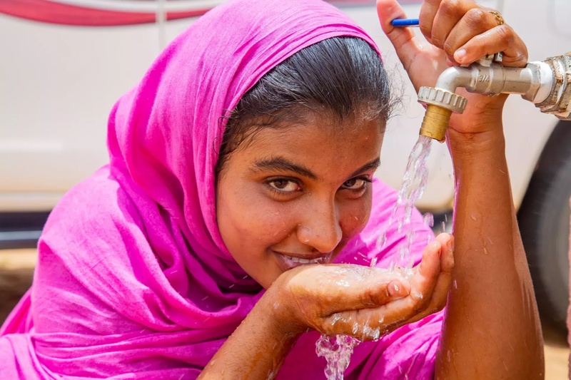 Une fille portant un foulard rose vif boit de l'eau à la main sous un robinet.