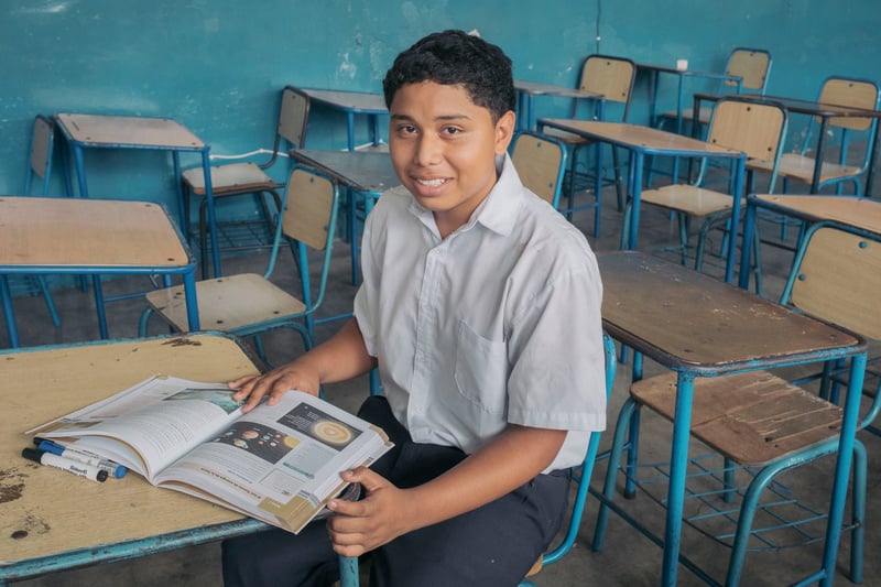 A schoolboy sits at a desk in a classroom with rows of empty desks behind him, a textbook showing planets open in front of him