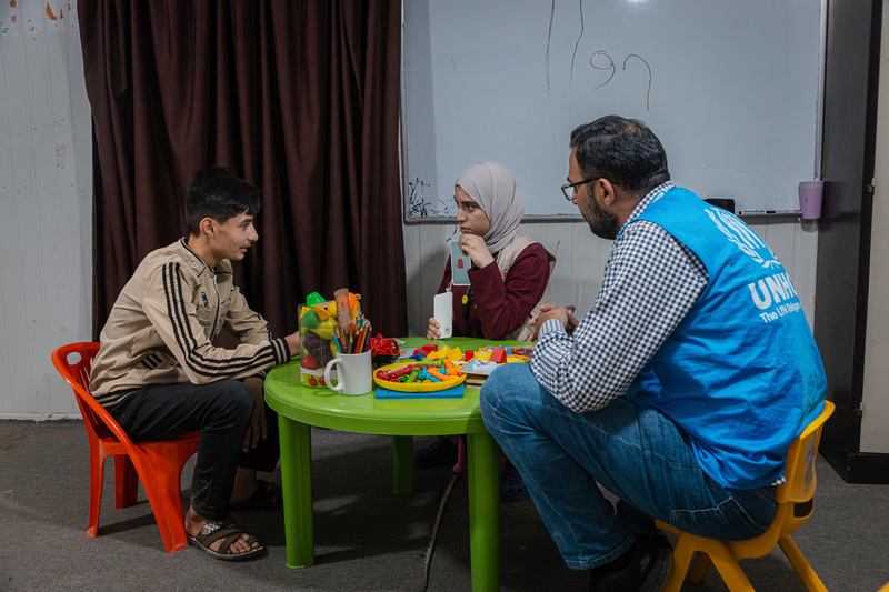 Seated on small plastic chairs at a green table, a young man speaks to a woman while a UNHCR staff member looks on.