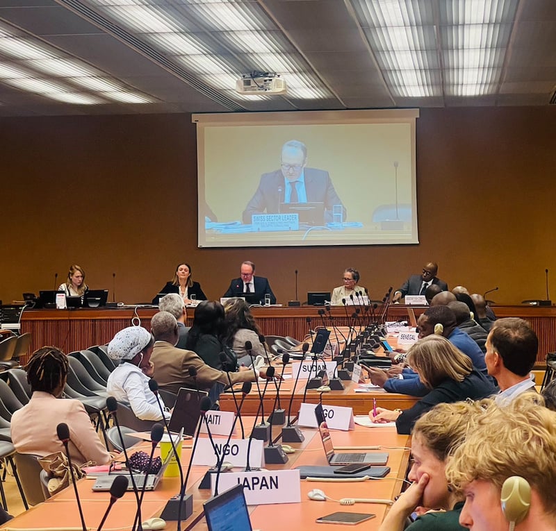 A panel of people sit at the top of a room, with a screen displaying a person above them. A table of participants sits in front, with microphones and laptops on the table.