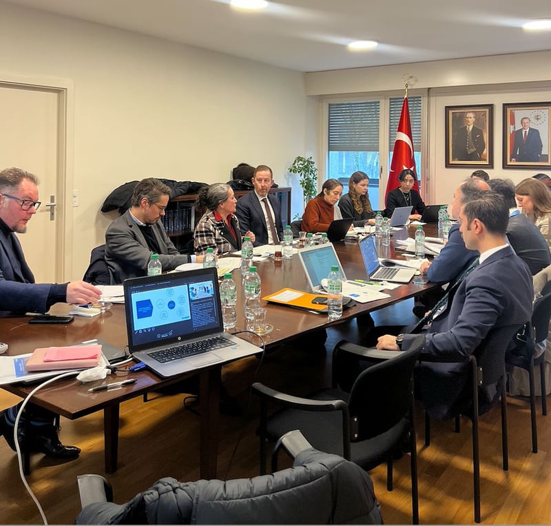 A group of 10+ people sit at a table in a meeting room, with laptops and water bottles on the table.