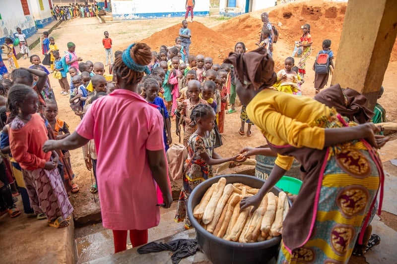 Women wearing aprons distribute bread from a large container to a group of children