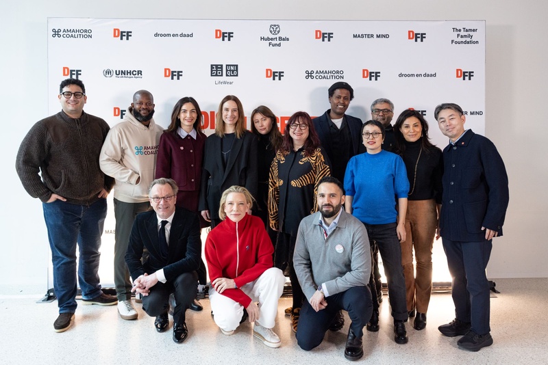 A large group of people smiling people pose for a photo in front of a large Displacement Film Fund banner featuring logos of many different organizations
