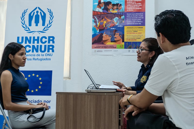 Sitting at a desk, a young woman is talking with a man and a woman who are on the other side of the desk