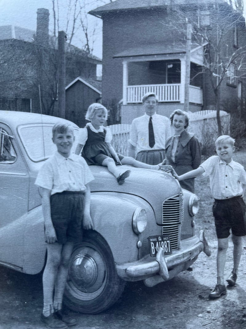 Une famille pose pour une photo en noir et blanc autour d’une voiture ancienne garée devant une maison