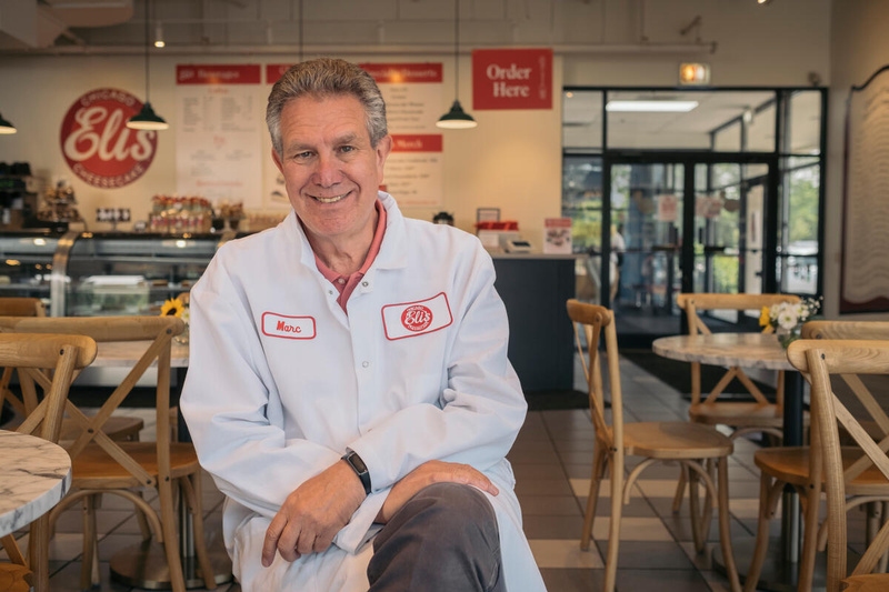 Marc Schulman, en blouse blanche de boulanger, est assis devant le comptoir d'un magasin de gâteaux au fromage.
