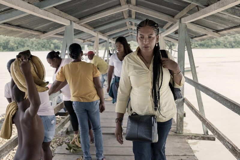 A woman walks past people on a covered wooden pier on a wide river.