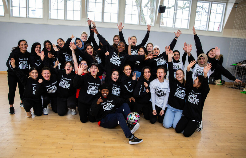 A group of around 30 young women in Girl Power sweaters smile with hands in the air
