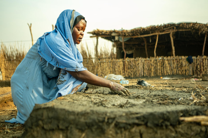A woman in a blue headscarf mixes mud and straw together to build a small structure.